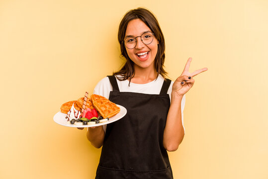 Young Hispanic Dependent Woman Holding Waffles Isolated On Yellow Background Joyful And Carefree Showing A Peace Symbol With Fingers.