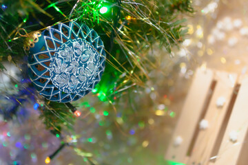 Closeup of the Christmas decoration (blue ceramic ball) with colorful garland on fir branch. Snowflakes on a wooden gift box near the tree. New year mood background