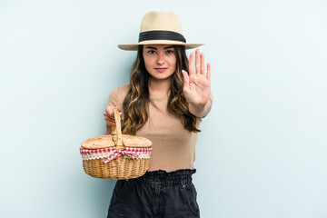 Young caucasian woman holding a picnic basket isolated on blue background standing with outstretched hand showing stop sign, preventing you.