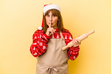 Middle age caucasian woman cooking cookies for christmas isolated on yellow background keeping a secret or asking for silence.