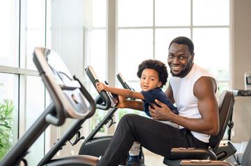 Fototapeta na wymiar African American man with child exercising on the exercise bike in indoor fitness club. Healthy and Fitness concept.