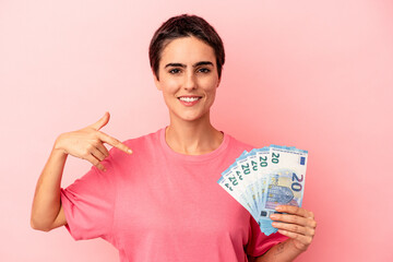 Young caucasian woman holding banknotes isolated on pink background person pointing by hand to a shirt copy space, proud and confident