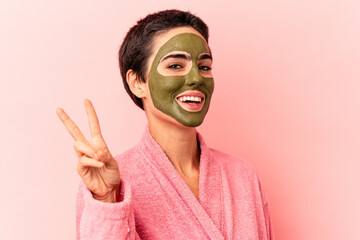 Young caucasian woman wearing a face mask isolated on pink background joyful and carefree showing a peace symbol with fingers.