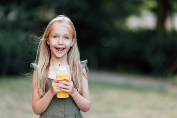 Candid lifestyle portrait of cute little caucasian girl with blonde hair and smiley face drinking fresh orange juice outdoor at hot summer day.