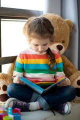A cute girl of 6 years old sits on the couch by the window with a large toy bear and reads a book. She is dressed in a colored jumper. Kids. Teaching.