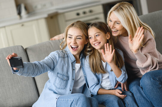 Three generations of Caucasian females. Happy grandmother, daughter and granddaughter, sitting on sofa in living room, having fun together, taking selfie posing for the camera, talking on a video call