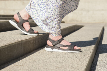 Womens legs in open leather sandals with flat soles go down the stairs. A woman in a light summer...