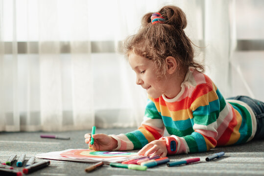 A cute little girl sits on the floor in the rays of the sun and draws a rainbow with colorful markers. She is dressed in a colored jumper. Creation. Childhood.