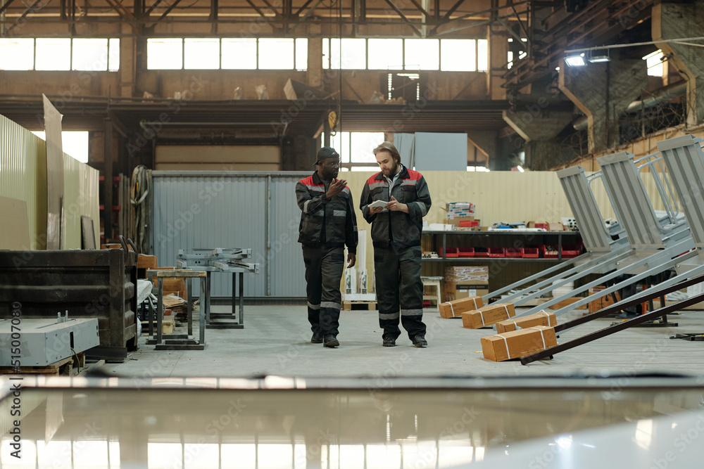 Poster two young intercultural male workers of factory having discussion of working points while moving alo