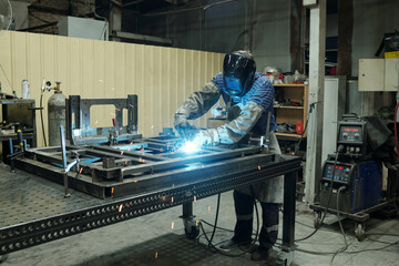 Young male worker of industrial plant bending over metallic workbench and welding parts of huge...