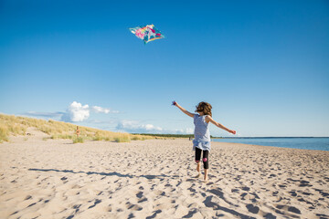 Cute happy little girl in summer dress running with flying kite on empty sandy beach. Beautiful sunny day, blue sky. - Powered by Adobe