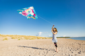 Cute happy little girl in summer dress running with flying kite on empty sandy beach. Beautiful sunny day, blue sky.