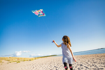 Cute happy little girl in summer dress running with flying kite on empty sandy beach. Beautiful sunny day, blue sky.