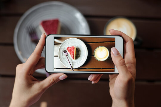 Hands of young female with smartphone taking photo of piece tasty cheesecake on saucer served with cappuccino in cafe