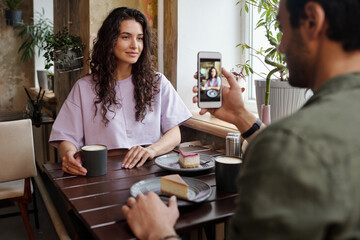 Young man taking photo of his girlfriend sitting by table in front of him and posing while having coffee with yummy dessert