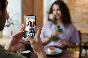 Hand of guy with smartphone taking photo of his girlfriend with rose and cheesecake sitting by table in front of him during romantic date