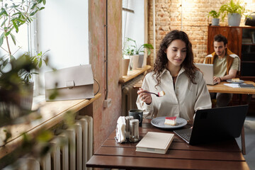 Fototapeta na wymiar Young businesswoman or freelancer in casualwear having dessert while sitting by table in front of laptop in cafe and networking