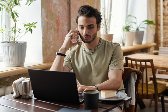 Young Male Freelancer In Casualwear Talking On Mobile Phone While Sitting In Front Of Laptop In Cafe, Networking And Having Coffee