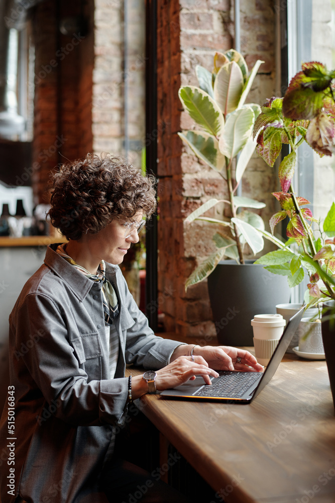 Poster Middle aged businesswoman or freelancer with laptop sitting in front of window in cafe and looking at screen while working over new project