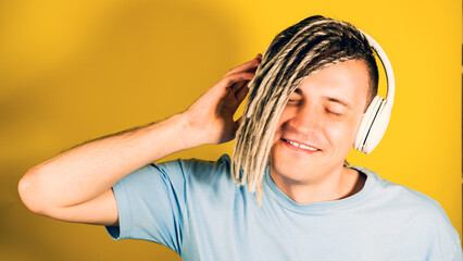 Young happy man in wireless white headphones listening to music on yellow background. Portrait of handsome smiling male enjoying songs with closed eyes.