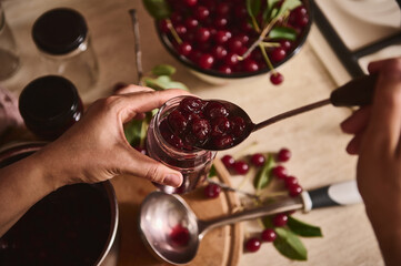 Housewife's hands filling a glass jar with homemade freshly brewed cherry jam over a pot of...
