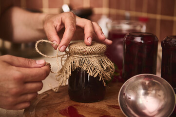 Hands of a housewife, chef confectioner tying bow with a rope around a burlap on a lid of a jar...