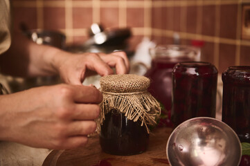 Housewife's hands putting burlap on the lid of a jar with freshly made jam and tying bow with a...