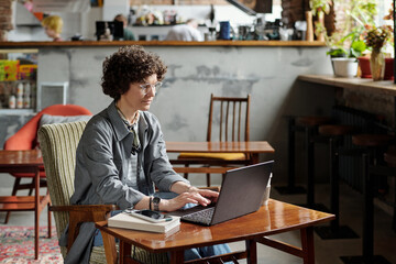 Obraz premium Young serious businesswoman in casualwear concentrating on organization of work while typing on laptop keyboard in cafe