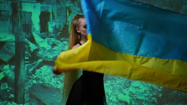 Woman shaking Ukrainian flag standing at background of ruins photo. Slim young sad lady with national flag at background of bombed building debris picture