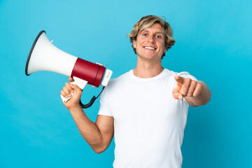English man over isolated blue background holding a megaphone and smiling while pointing to the...