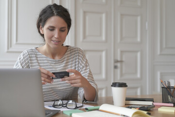 Portrait of cheerful businesswoman sitting at desk in front of laptop while looks at credit card