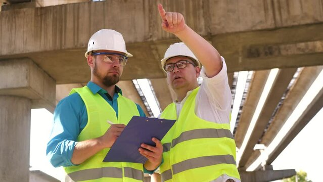 Two civil engineers working on a project wearing safety helmets are discussing real estate development and planning at a construction site together. The foreman and his assistant are talking to each
