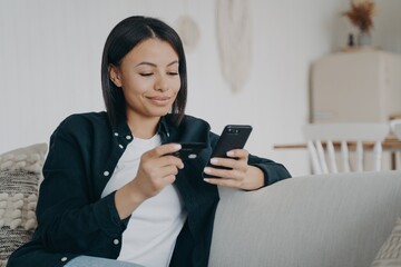Fototapeta premium Hispanic girl is shopping online. Young happy woman is holding smartphone and credit card.