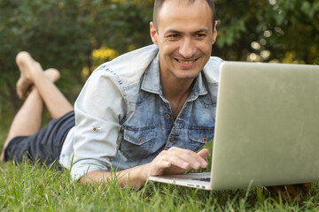 Freelancer man with laptop working in garden sit in chair on grass outdoors. Young blogger male work on computer in public park processes video for social media content. Place of work, distance job.