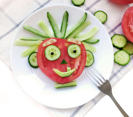 Kids food. Funny cheerful face made of tomato and cucumbers on a plate close-up on a white...