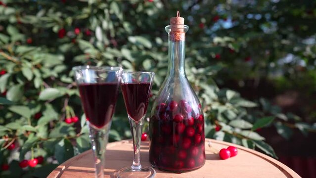 Homemade cherry brandy in two glasses and in a glass bottle on a wooden table in a summer garden, close up, rotates