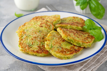Zucchini fritters with cheese, garlic and herbs. Vegetarian zucchini pancakes, served with basil and sour cream on a gray concrete background. Selective focus.