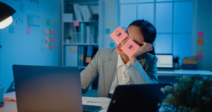 Young Asian businesswoman sitting on desk overworked tired sleep over a laptop at office at night. Exhausted burnout lady with two post-it over her eyes, adhesive notes on face sleeping at workplace.