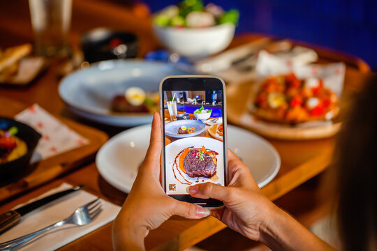 Woman makes mobile phone photo of baked beef meat in luxury restaurant. Smartphone food photography of fine food dinner. Woman hands taking phone photo of food in trendy style for social media