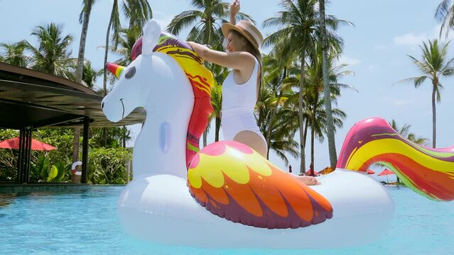 Summer vacation. Beautiful slim young woman in swimwear enjoying time in swimming pool floating in a big inflatable unicorn toy on sparkling blue pool, dancing and celebrating holidays in Thailand