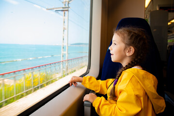 A little girl of 6-7 years old looks out the window of a train at the sea. She's wearing a yellow parka. Journey. Reflection. Vacation. Summer. Family vacation.