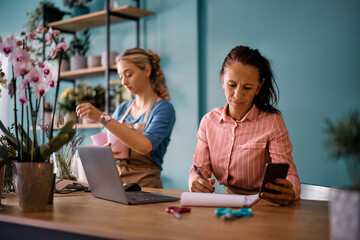 Flower shop owner using cell phone while writing orders on paper at her store.