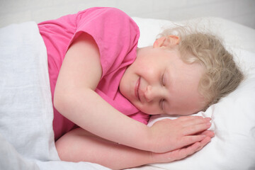 cute little girl with blond hair and a pink t-shirt sleeps on a white bed