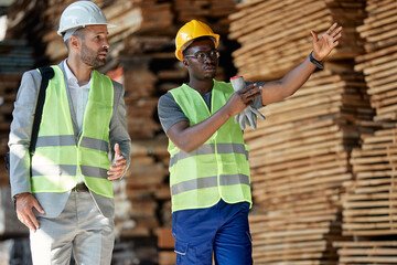 African American worker talks to inspector while guiding him through wood warehouse.