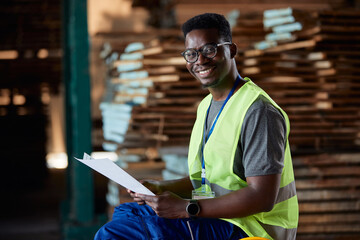 Happy black worker going through reports at wood warehouse and looking at camera.