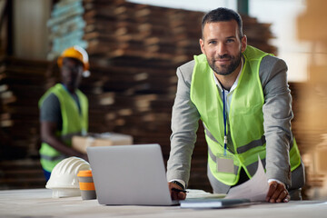 Happy businessman working on laptop at wood warehouse and looking at camera.
