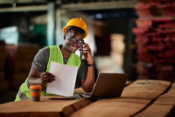 Happy black worker talking on cell phone while reading reports and using laptop at wood warehouse.