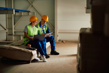Warehouse workers collaborate while using laptop and going through paperwork in storage room.