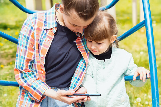 A Teenage Girl Teaches Her Younger Sister How To Use A Cell Phone. Happy Sisters Play Together On A Smartphone. A Young Babysitter Shows A Little Girl A Video On A Smartphone Screen.Mobile Phone Games