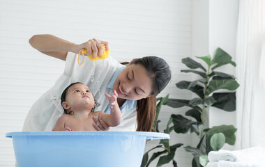 Asian little baby bathing in bathtub and looking at sponge which young mother squeeze water out...